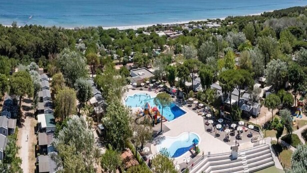An aerial view of a coastal resort with a central swimming pool, surrounded by dense greenery and beach access.