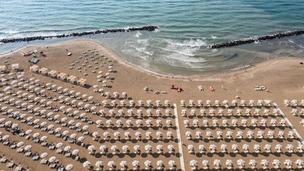 An aerial view of a beach with rows of umbrellas and sun loungers, a rocky breakwater, and calm sea. - Lodge in Rimini Family Village near the beach (Viserba, Rimini)