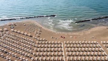 Vue aérienne d’une plage bordée de rangées de parasols et de chaises longues, d’une digue rocheuse et d’une mer calme.
