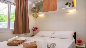 A hotel room with a bed, towels, a nightstand, a plant, and a window with curtains. - Lodge in Rimini Family Village near the beach (Viserba, Rimini)