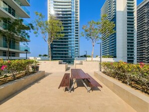 A rooftop terrace with a wooden table and benches, surrounded by low-rise buildings and greenery. - Lux 3 Bed | Private Beach Access, Emaar Beachfront I Sunrise Bay Tower 2 (Emaar Beachfront)