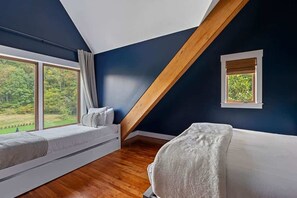 Ein Schlafzimmer mit schrägverdeckter Decke, einem Fenster mit Blick auf Grünflächen, einem Bett mit weißer Bettdecke und einem kleinen Nachttisch.