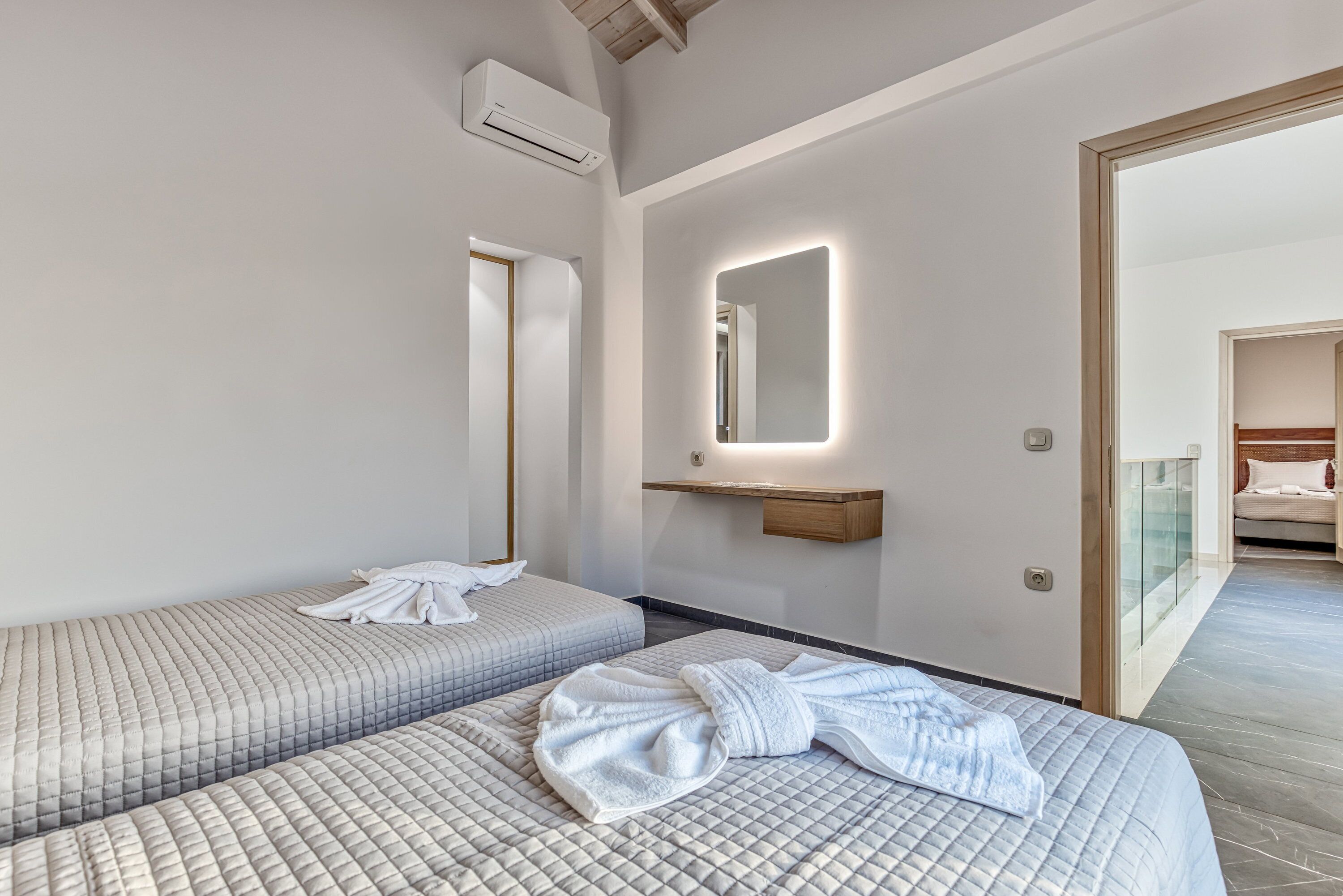 Ein Hotelzimmer mit zwei Betten, einem beleuchteten Spiegel, einem Holzregal und einer Glastür zu einem anderen Raum.