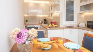 A modern kitchen with white cabinets, a dining table set with plates and a vase of flowers, and a fruit bowl on the counter. - Bethy's #1|Double BR Apt (Corfu)
