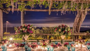 An outdoor dining setup with tables set for a meal, surrounded by greenery and flowers, with a cityscape in the background. - Villa Sogno by My Villa Collection (Sant'Agnello di Sorrento)