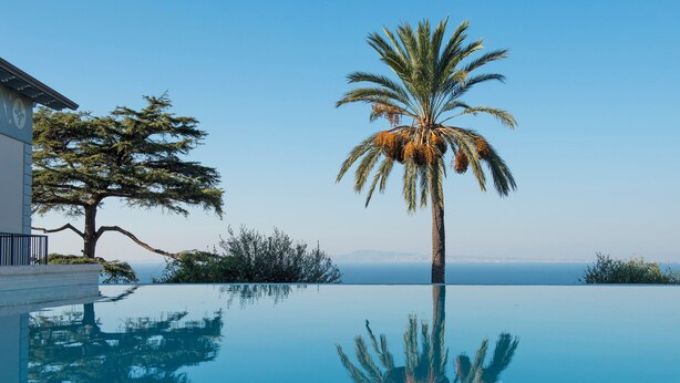 A clear pool reflecting palm trees and a building, with a view of the sea and mountains in the background. - Villa Sogno by My Villa Collection (Sant'Agnello di Sorrento)