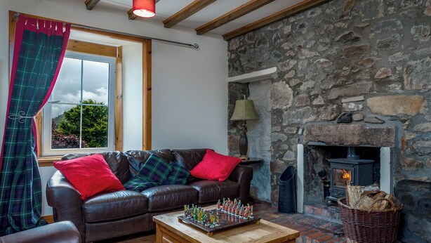 Una sala de estar acogedora con chimenea de piedra, un sofá de cuero con cojines de cuadros, una mesa de centro de madera y una ventana con cortinas.