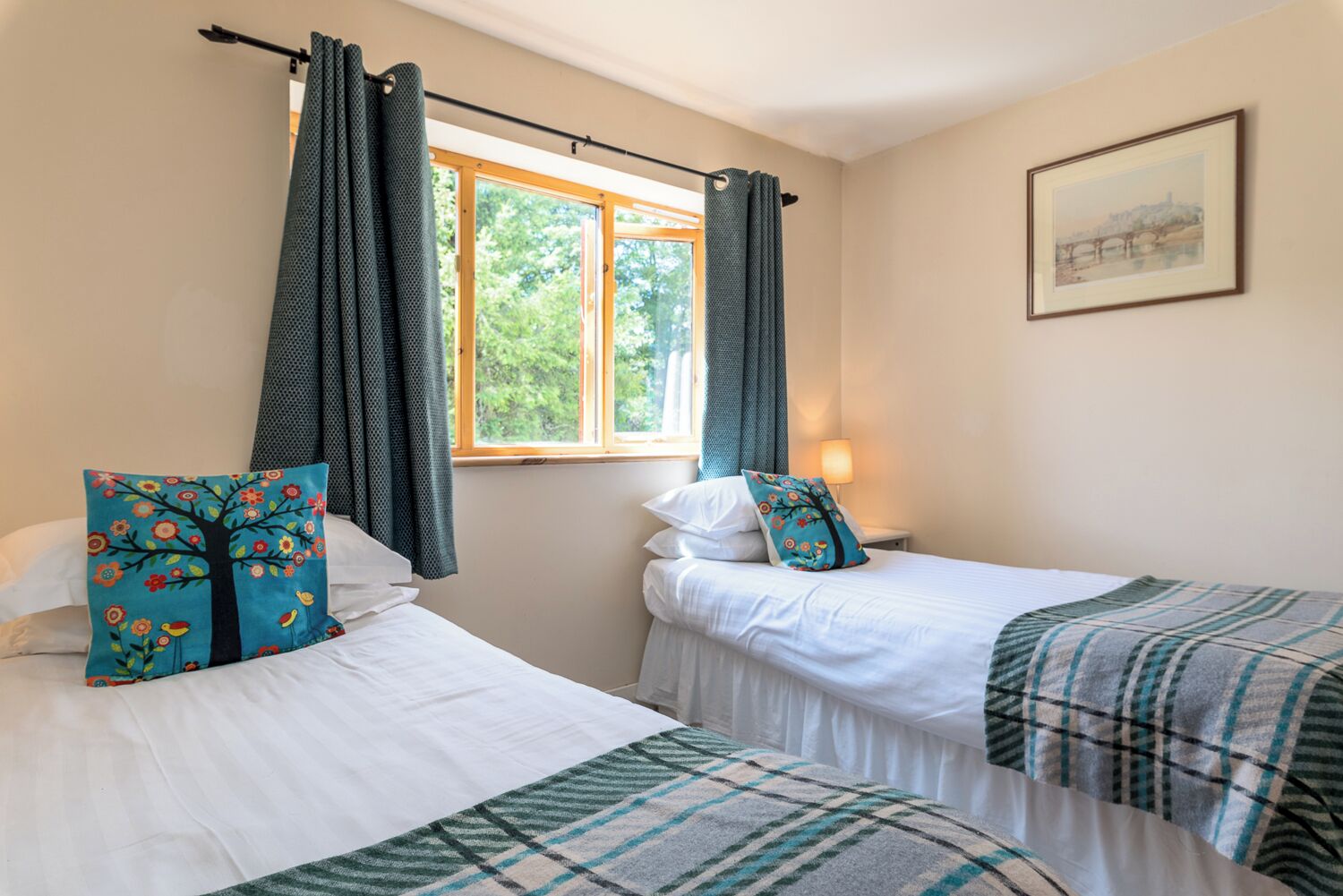 Una habitación con dos camas individuales, cada una con almohada y manta. Se ve una ventana con cortinas, una lámpara en una mesita de noche y un cuadro en la pared.