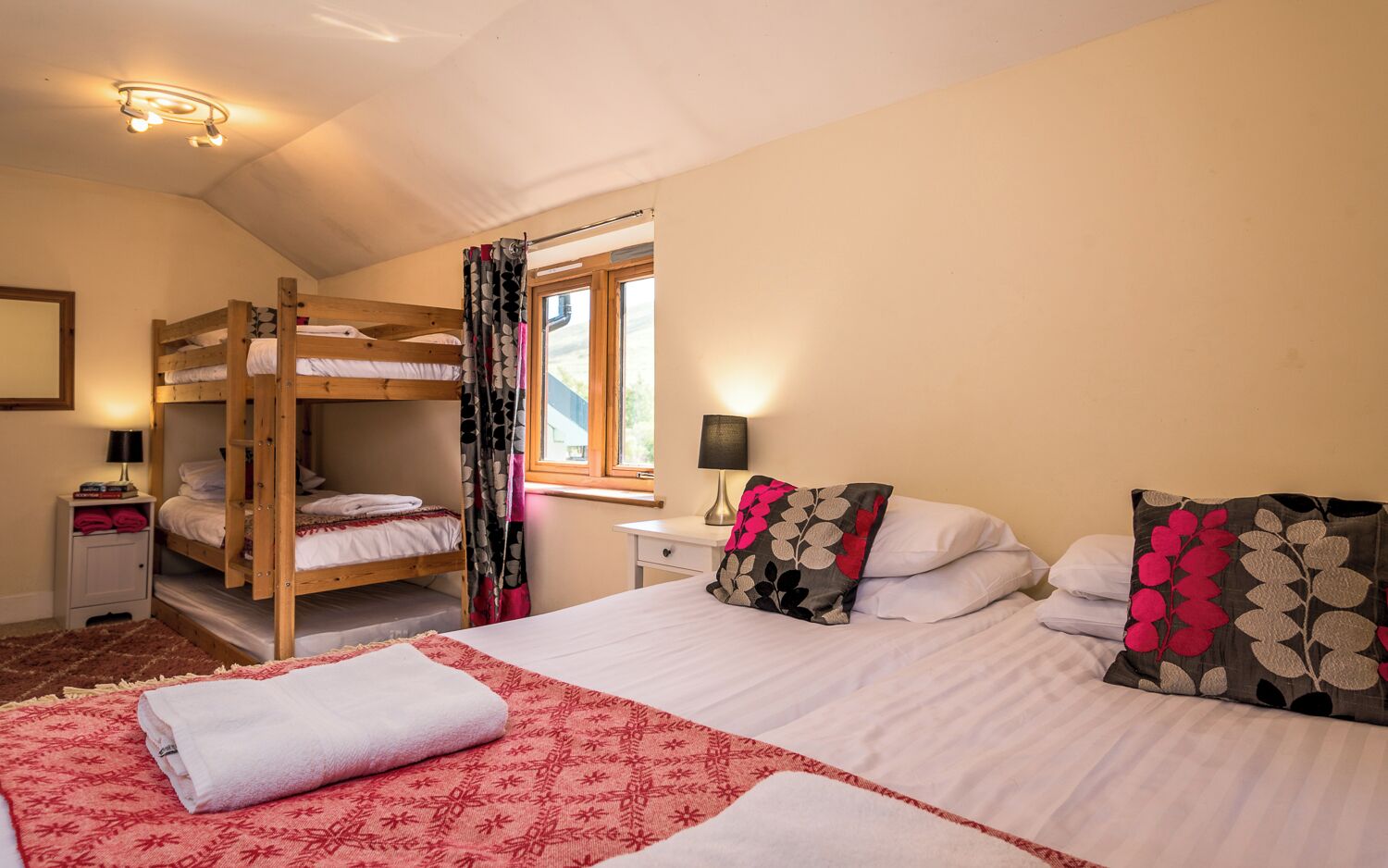 Un dormitorio con literas, una cama grande con almohadas, una ventana con cortinas, una mesita de noche con lámpara y un mueble pequeño.