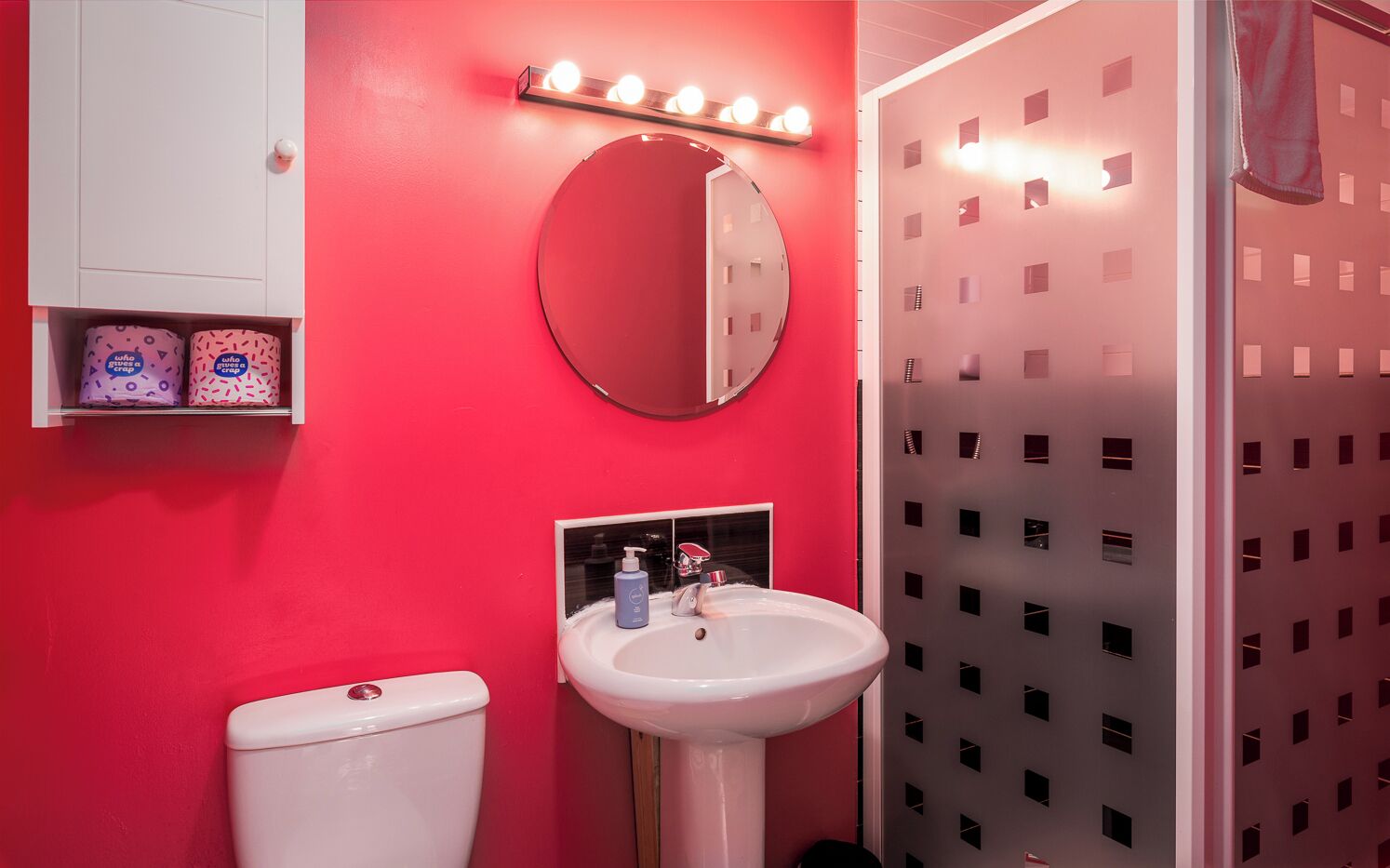A bathroom with a red wall, a white sink, a toilet, and a mirror with lights above it.