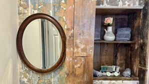 Une salle de bain rustique avec un meuble-lavabo en bois, un miroir rond, un vase contenant une seule fleur et des articles de toilette sur l’étagère.