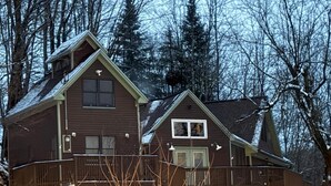 Una casa de madera de dos pisos con balcón, rodeada de nieve y árboles.