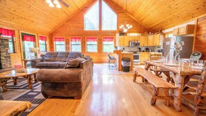 A cozy cabin interior with a wooden ceiling, a living area with a sofa and coffee table, a kitchen with wooden cabinets, and a dining table.
