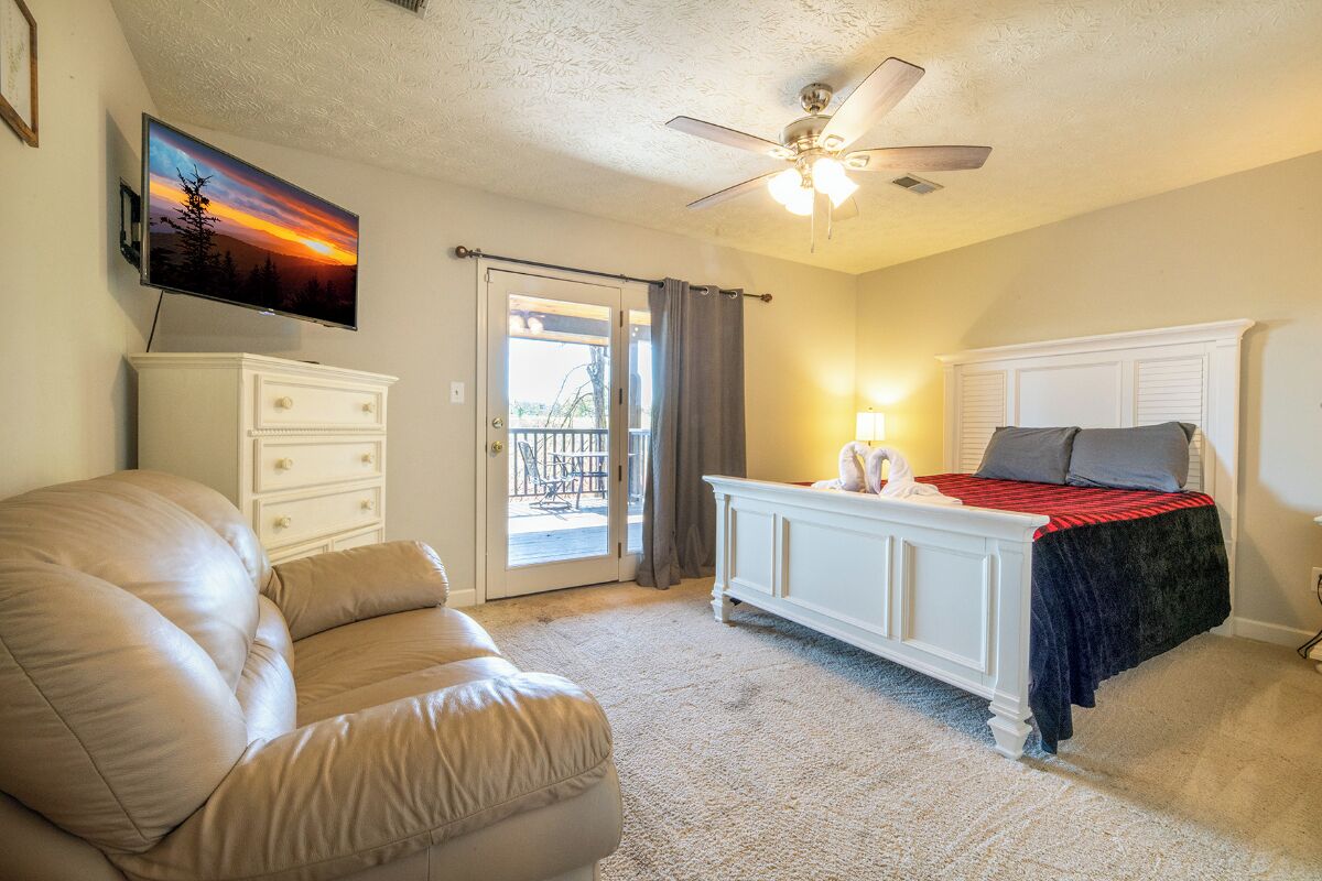 A bedroom with a bed, a white dresser, a beige sofa, a ceiling fan, and a TV mounted on the wall.