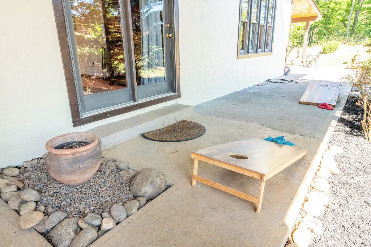 A patio with a concrete floor, a small wooden table, a potted plant, and a window with a view of trees.