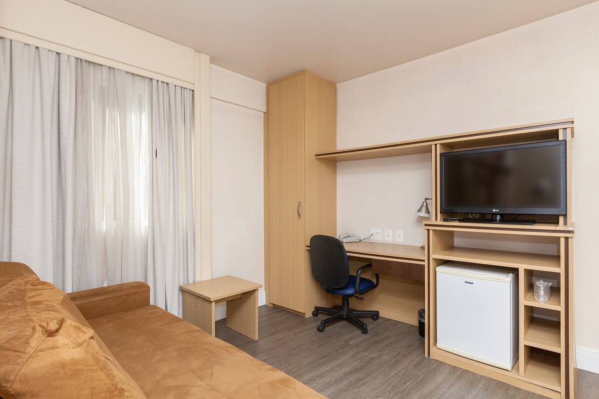 Habitación de hotel pequeña con cama, escritorio con computadora, televisor y refrigerador miniatura.