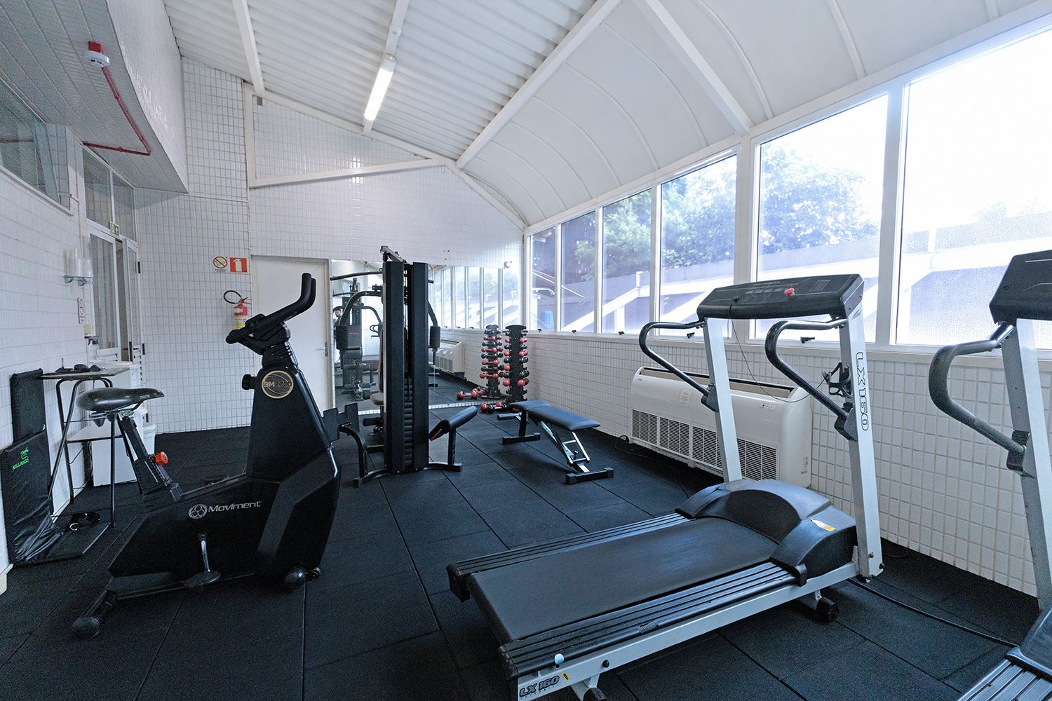 Un gimnasio con diversos equipos de ejercicio, incluyendo cintas de correr y máquinas de pesas, con amplias ventanas que permiten la entrada de luz natural.