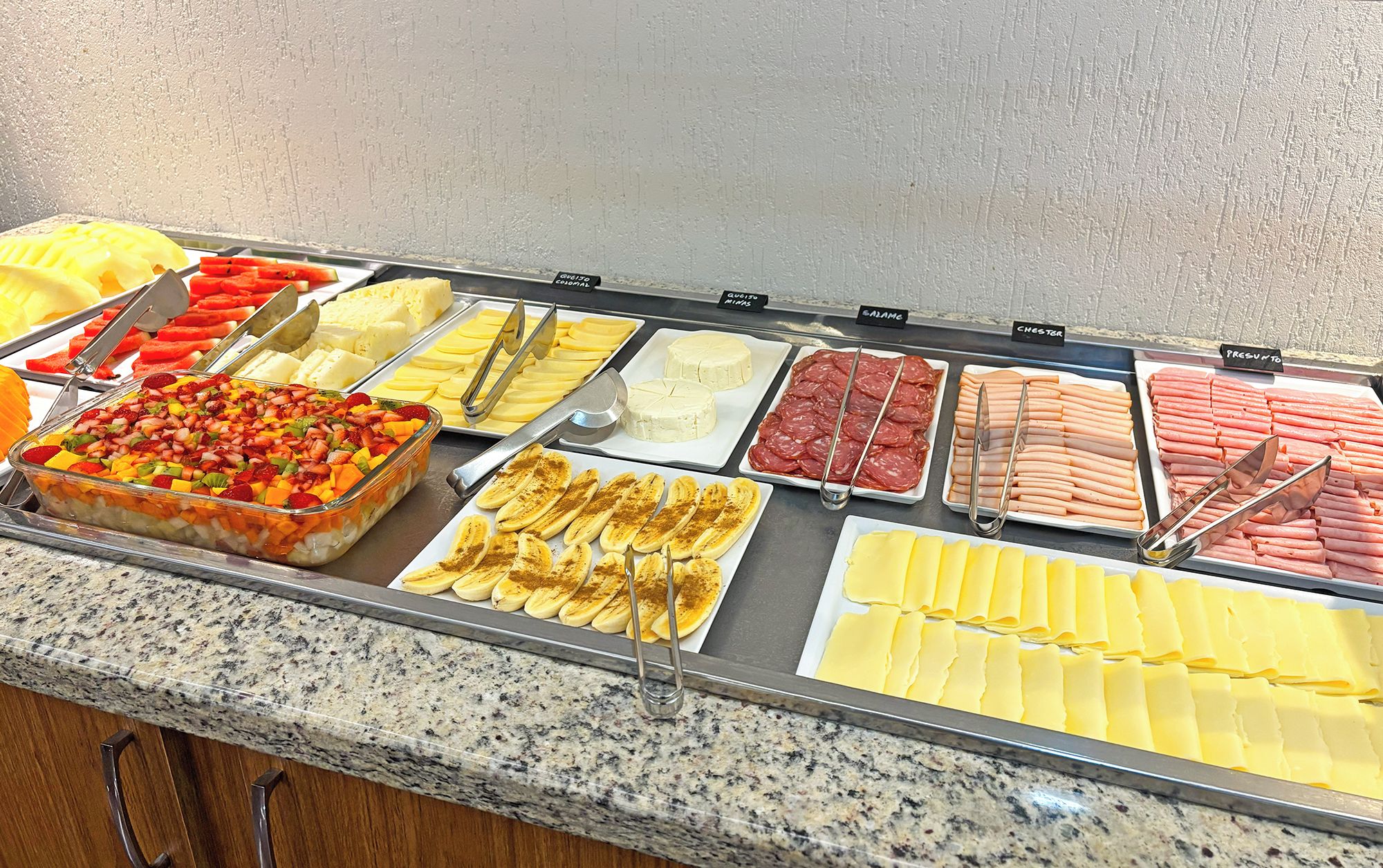 A buffet with various food options including fruits, cheeses, meats, and a fruit salad.