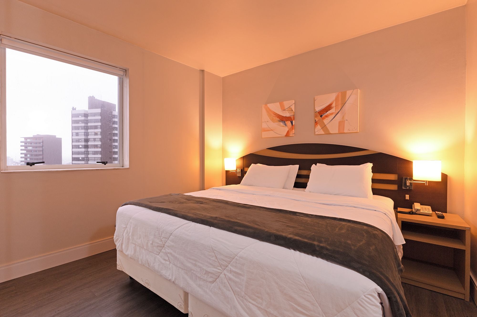 A hotel room with a large bed, bedside tables, a window with a view of buildings, and abstract wall art.