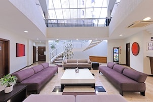 Un vestíbulo espacioso con techo alto, un área de estar central con sofás, una recepción y una escalera que conduce a un nivel superior.