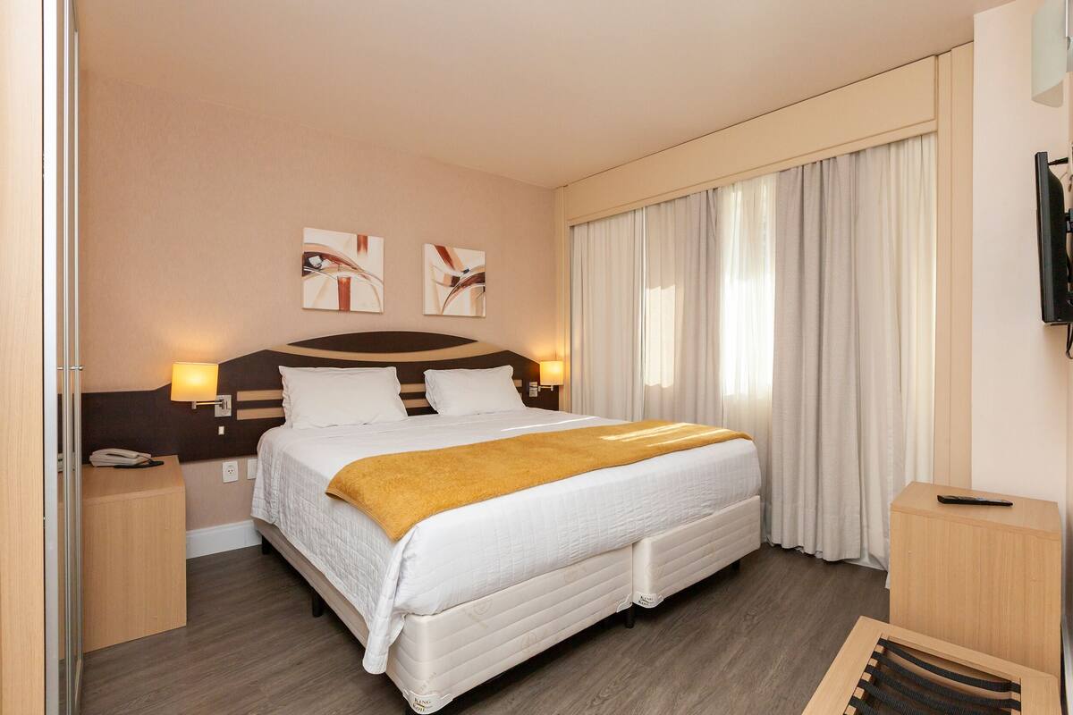 Habitación de hotel con una cama grande, una mesita de noche, un televisor y una ventana con cortinas.