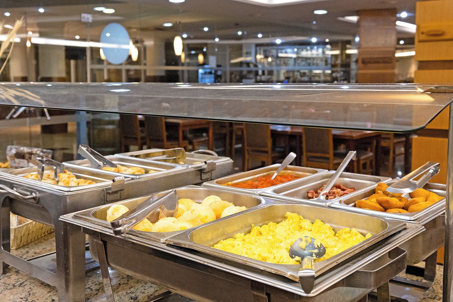 A buffet with various food items in metal containers, a glass barrier, and a modern interior with a ceiling light.