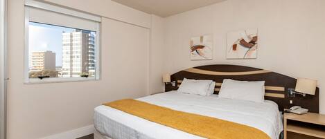 Une chambre d’hôtel avec un lit, une fenêtre donnant sur des immeubles et deux lampes fixées au mur.