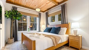 A bedroom with a wooden bed, a nightstand, a lamp, and a large window with curtains. - Luxe Retreat: Sleeps 16, Hot Tub, Sauna & Movie Room (Bethel)