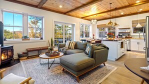 A cozy living room with a sectional sofa, a fireplace, and a large window overlooking a forest. - Luxe Retreat: Sleeps 16, Hot Tub, Sauna & Movie Room (Bethel)