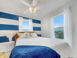 Un dormitorio con cama, mesita de noche, lámpara, ventilador de techo y ventana con cortinas.