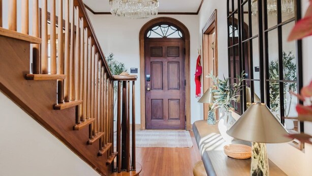 A wooden staircase with a chandelier, a wooden door, and a table with a lamp and decorative items. - Hoosier Memories! Luxury Stay, Steps from IU - Pool Table, Firepit & More (Bloomington)