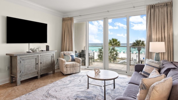 A living room with a flat-screen TV, a sofa, a coffee table, a chair, a cabinet, and a view of the beach.