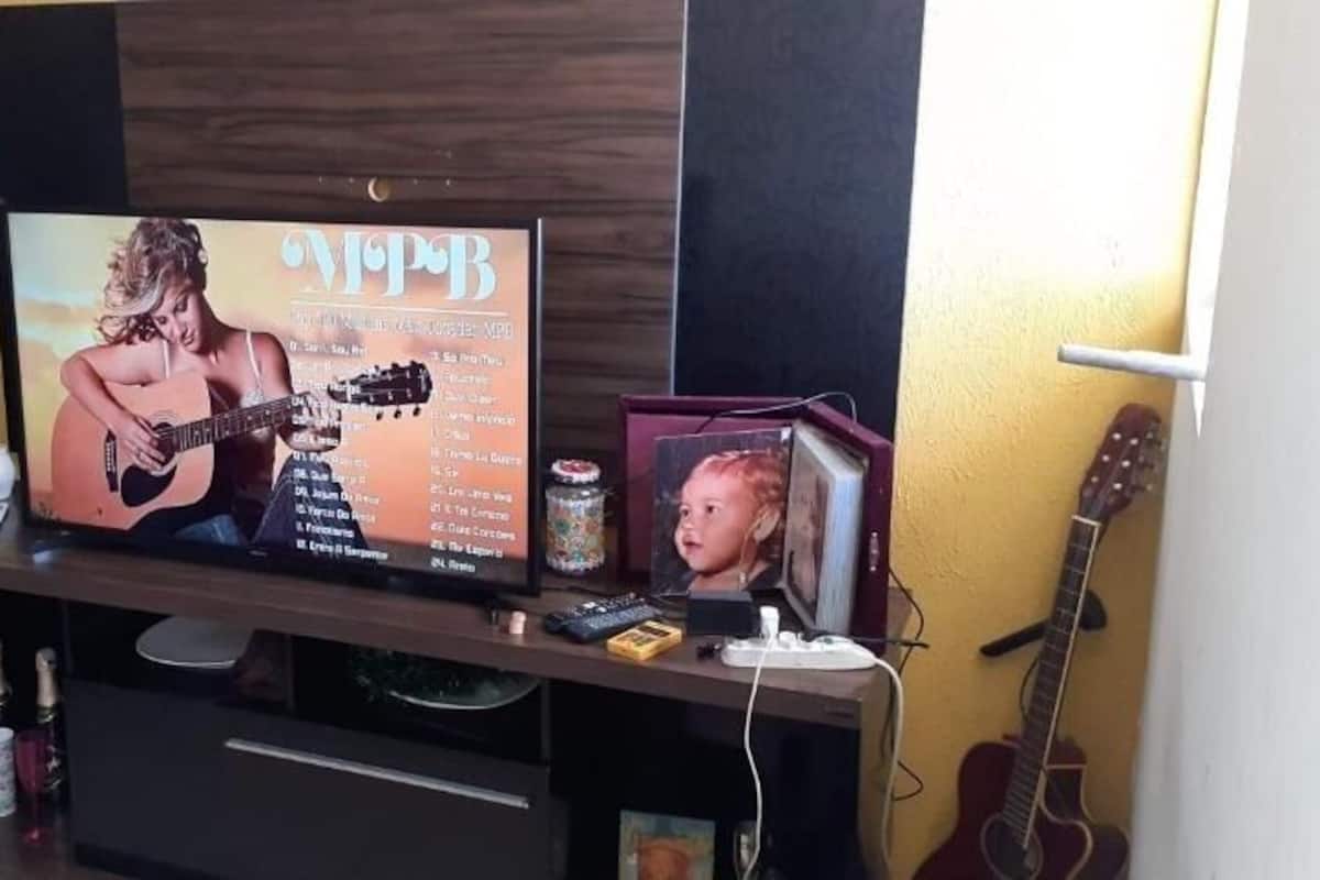 Ein Wohnzimmer mit Fernseher, der ein Albumcover zeigt, einer Gitarre, einem kleinen Tisch mit einer Vase und einem gerahmten Bild.