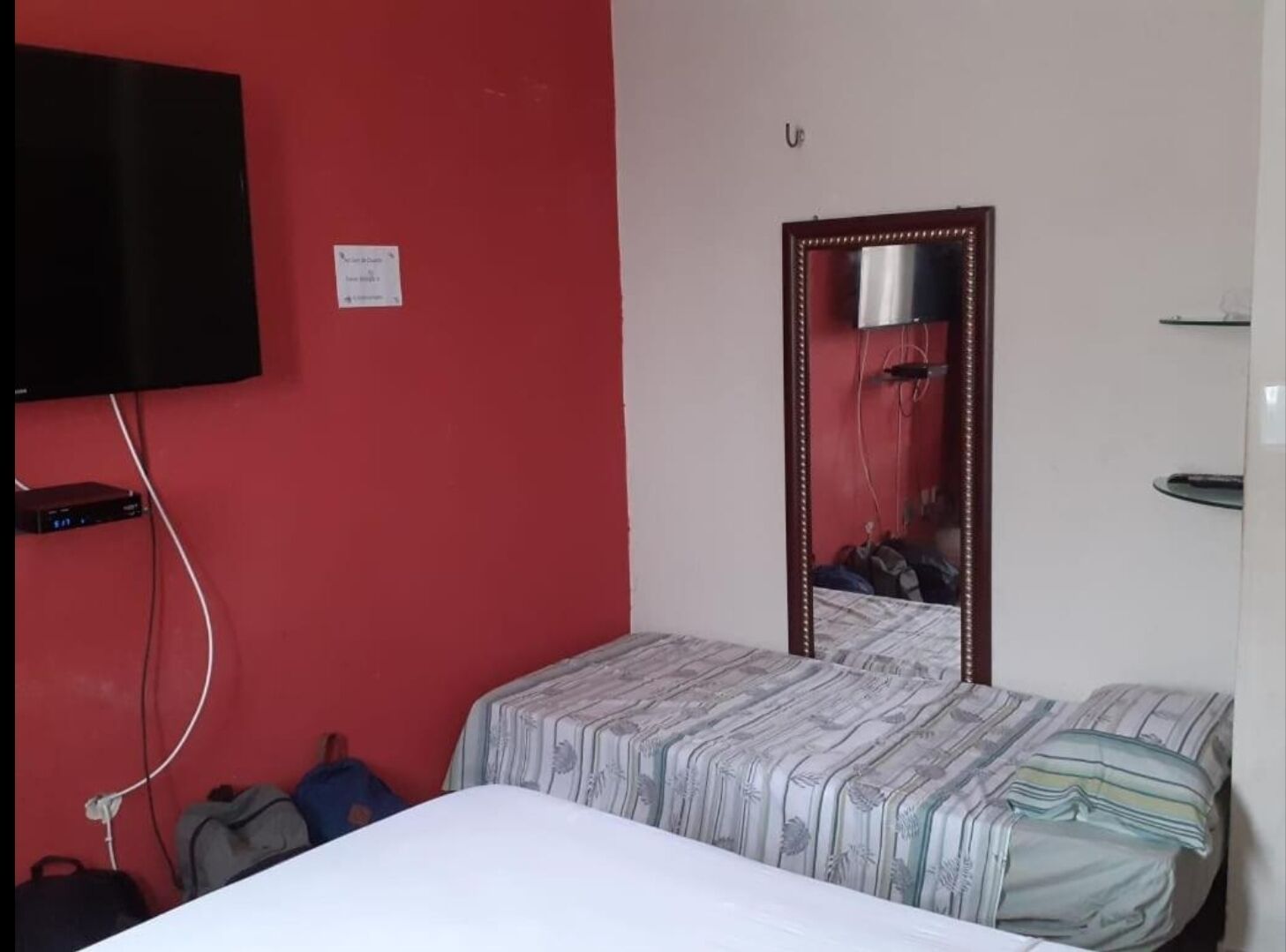 A small, simple room with a bed, a mirror, a TV, and a red wall.