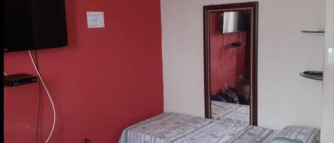 A small, simple room with a bed, a mirror, a TV, and a red wall.