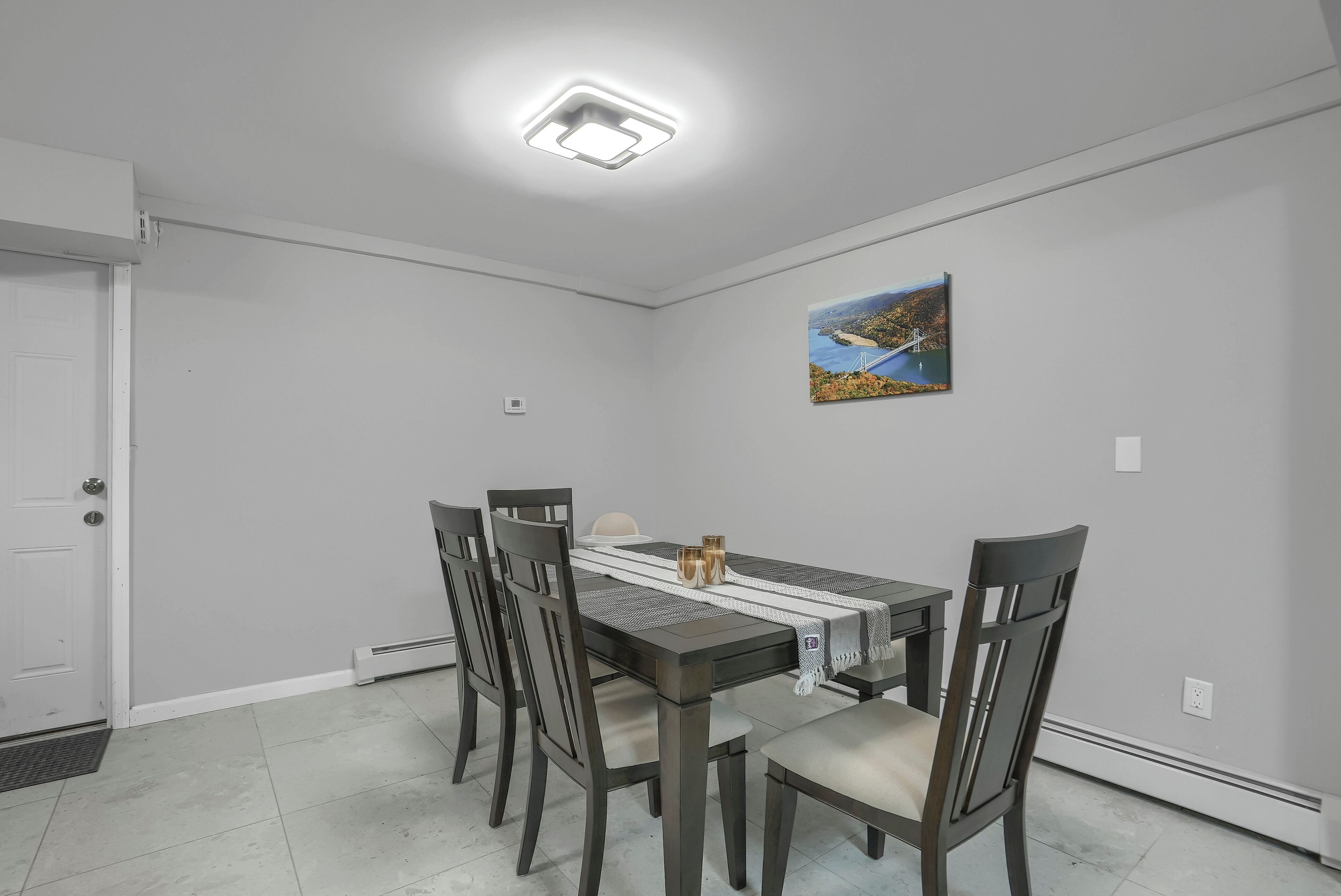 A dining room with a rectangular table, four chairs, a white door, a ceiling light, and a framed picture on the wall.