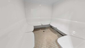 Une salle de bain aux murs et au sol recouverts de carreaux blancs, avec trois lavabos blancs et un drain au centre.
