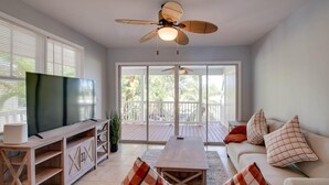 A spacious living room with a large flat-screen TV, a wooden entertainment unit, a white sofa with plaid cushions, a wooden coffee table, and a ceiling fan. - Cabana at the Golf Club-Monthly Rental Charming 2- (Key West)