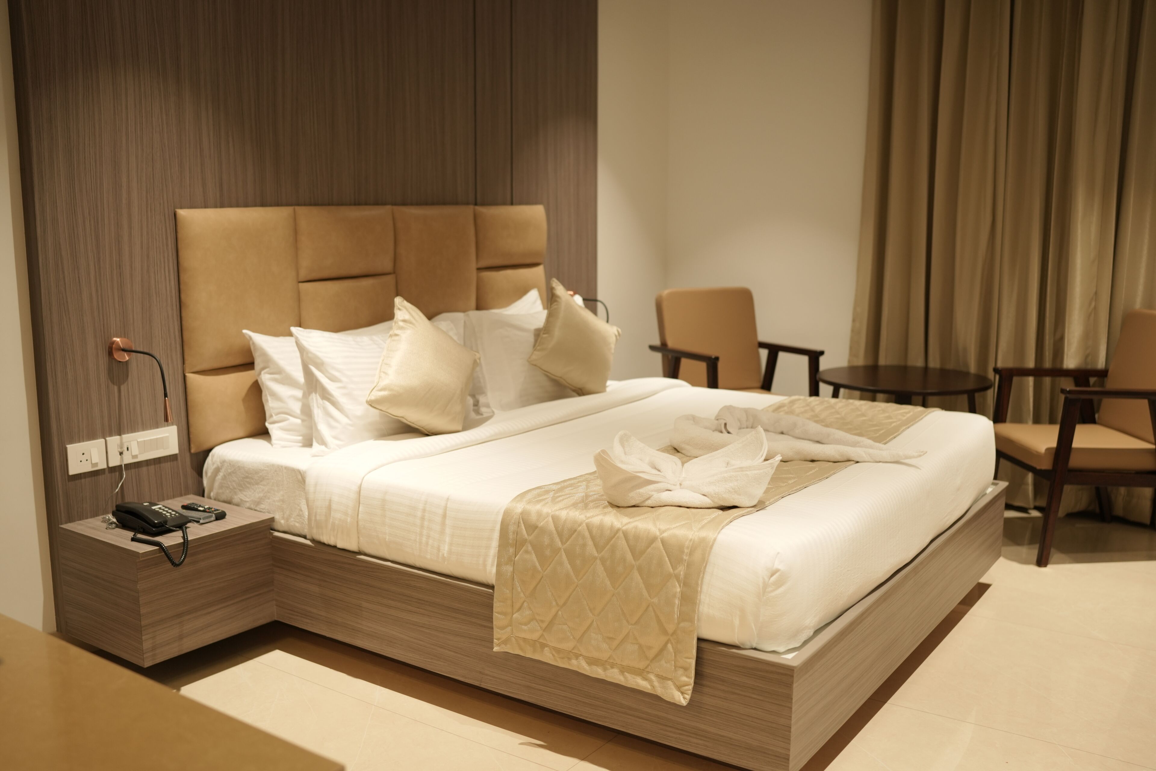 A modern hotel room with a large bed, a bedside table with a phone, a small table, and a chair.
