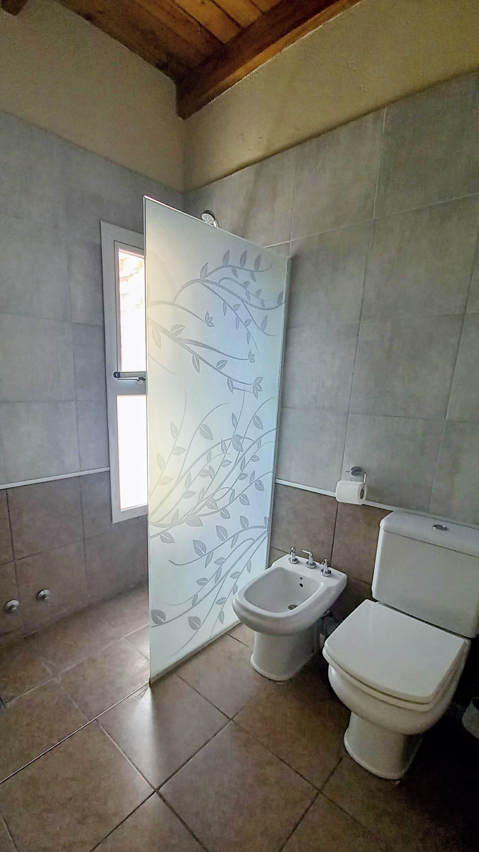 A bathroom with a bidet, toilet, and a frosted glass shower door with a floral design.