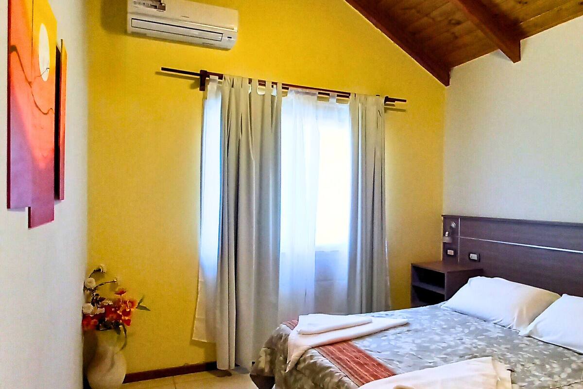 Egy hálószoba, amelyben található egy ágy, egy mennyezeti ventilátor, ablakfüggönnyel ellátott ablak és egy légkondicionáló berendezés.
