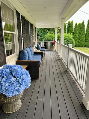 A covered porch with a seating area, a wicker basket with blue flowers, and a view of a green landscape.