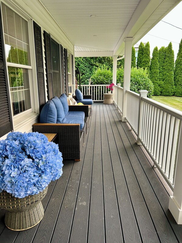 A covered porch with a seating area, a wicker basket with blue flowers, and a view of a green landscape. - Spacious 5bdrm home- saltwater pool, WiFi, AC, private beach parking pass (Westerly)