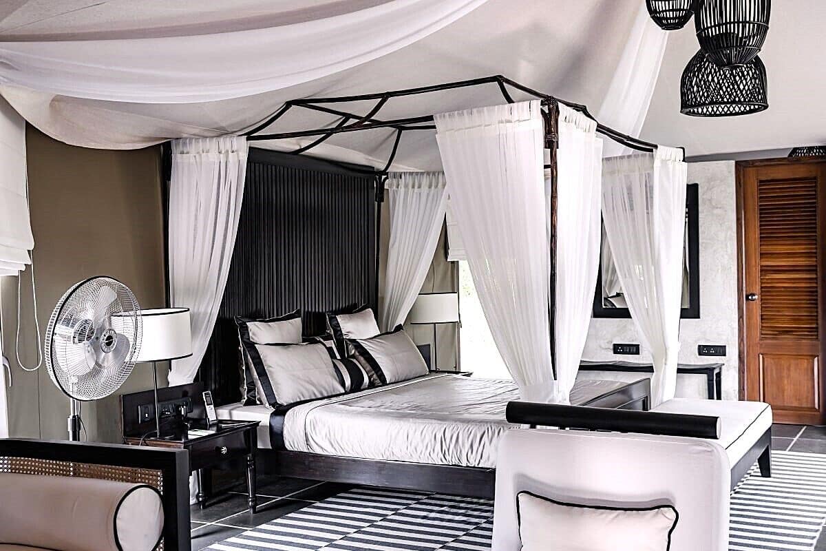 A modern bedroom with a four-poster bed, a bedside table, a chair, and a fan.