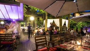 An outdoor dining area with wooden tables and chairs, covered by umbrellas, and a stage with a light display. - Hotel Milano Pool & Garden (Godiasco)