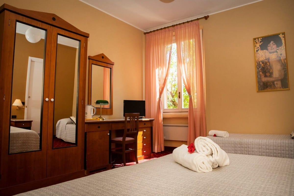 A hotel room with a wooden wardrobe, a desk with a computer, a bed with a rolled towel, and a window with curtains.