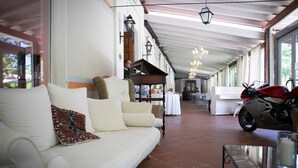 A spacious living area with a white sofa, a motorcycle, and a dining area in the background. - Hotel Milano Pool & Garden (Godiasco)