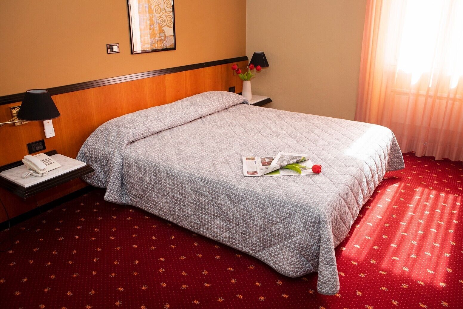 A hotel room with a bed, a nightstand, a lamp, a telephone, and a vase with flowers.