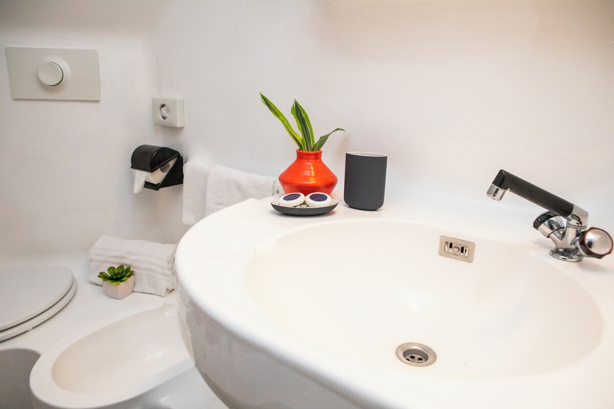 A bathroom with a white sink, a toilet, a bidet, a red vase with a plant, and a black cup.