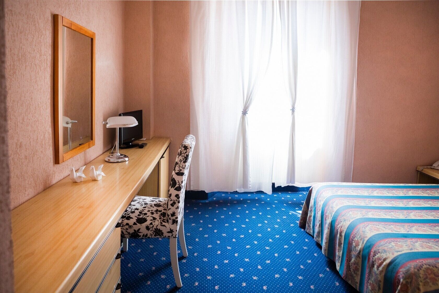 Quarto de hotel com escrivaninha de madeira, uma cadeira, cama com roupa de cama estampada e uma janela grande com cortinas leves.